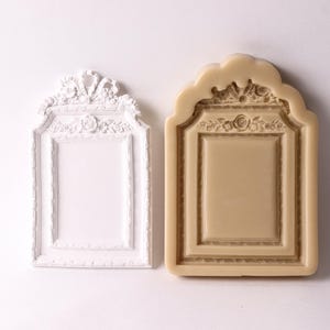 May include: A white ornate picture frame mold and a brown silicone mold of the same design. Both molds are rectangular with a decorative top and bottom.