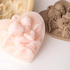 May include: A light pink heart-shaped casting featuring two cherubs in relief, a beige silicone mould, and a brown heart-shaped casting with a detailed cherub design. Each piece displays a small hole at the top for hanging or attachment.