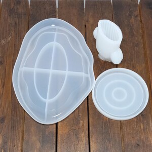 May include: Three white silicone molds for resin crafts. The largest mold is a freeform oval shape with a divided interior. The second mold is a round shape with concentric circles. The third mold is a small, freeform shape with a cylindrical opening.