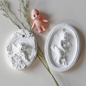 May include: Two white ceramic ornaments featuring angel figures. One ornament depicts a sleeping angel with wings and a floral design. The other ornament depicts a standing angel with wings and a floral design.