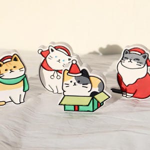 May include: Four adorable cartoon cats wearing Santa hats. One cat is holding a gift box, another is wearing a green scarf, and two are wearing red Santa suits.