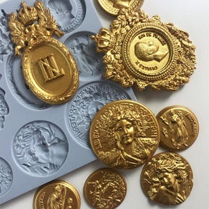 May include: A set of gold-colored metal molds for making decorative ornaments. The molds feature various designs, including a crown, a shield with a royal crest, and a portrait of a woman. The molds are made of silicone and are flexible and easy to use.