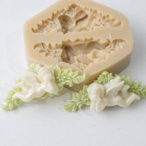 May include: A beige silicone mould featuring two recessed cavities shaped like cherubs surrounded by leafy foliage. Two finished light green and cream-coloured cherub figures with leaf accents are positioned in front of the mould.