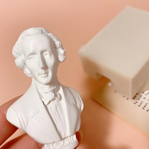 以下が含まれることがあります： ベースに「CHOPIN」という名前が入った、フレデリック・ショパンの白い胸像。胸像の隣には、印刷されたテキストが書かれた本のページの上に、2つの長方形のオフホワイトのバー（おそらく石鹸）があります。背景はソフトピーチ色です。