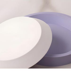 May include: A two-piece container with a lid. The lid is white and round, and the base is a light purple color. The container is likely used for storage or display purposes. The design is simple and modern.