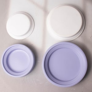 May include: Four round, flat, decorative plates. Two are white and two are a light purple.