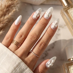 Peut inclure: Une main avec de longs ongles blancs et scintillants, chacun orné d'un motif de croix blanche. La main porte également deux bagues dorées et une alliance sertie de diamants. Les ongles sont en forme d'amande.