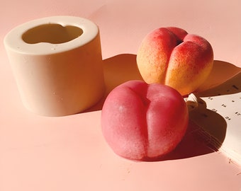 May include: A cylindrical off-white silicone mold sits next to a realistic peach-shaped candle with a yellow and red gradient and a second solid dark pink peach-shaped candle. The items are arranged on a light pink surface with a partial calendar.