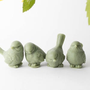 May include: Four small green ceramic bird figurines. The birds are in various poses, with some looking down and others looking straight ahead.