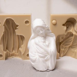 May include: A white figurine of a person holding a small child, positioned in front of two tan silicone moulds. The moulds feature a recessed, detailed impression of the same figure, with circular alignment points visible on the rectangular blocks.