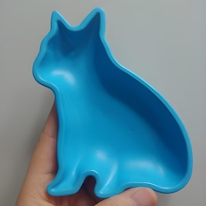 May include: A bright blue cat-shaped dish. The dish is made of a smooth, solid material and is designed in the shape of a sitting cat. The dish has a curved, open top, and is held by a hand against a neutral background.