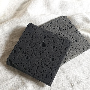 May include: Two square, porous coasters. One is black, the other is gray. Both have a rough, textured surface with small holes. They are displayed on a white, textured cloth. These coasters are designed to absorb moisture.