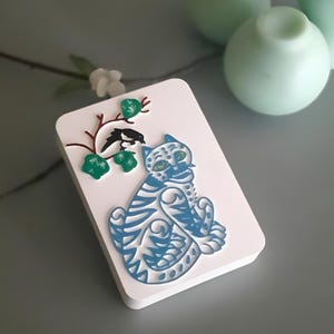 May include: A white rectangular decorative piece with a blue cat design. The cat is stylized with intricate patterns. A branch with green leaves and a black bird is on the left side. The background is a light gray.