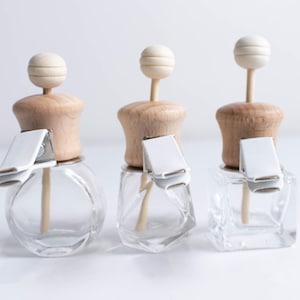 May include: Three glass bottles with wooden tops and white clips. Each bottle has a wooden stick with a white ball on top. The bottles are clear and have a round, square, and rectangular shape.