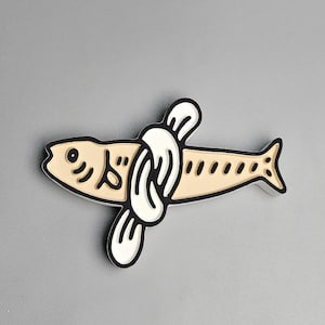 May include: A beige fish-shaped pin with black outlines and a white knot design. The pin has a cartoonish style with a small eye and dotted details. The background is a solid gray.