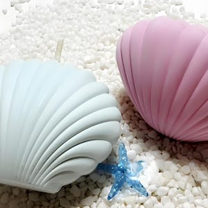May include: Two seashell-shaped candles, one light blue and one pink, rest on a bed of white pebbles. A small blue starfish is placed between the candles. The candles have a ribbed texture, and the scene evokes a beach theme.