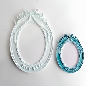 May include: Two oval-shaped picture frames with ornate details. The larger frame is white with a light blue wash and the smaller frame is teal blue.