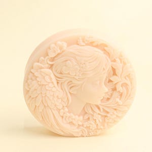 May include: A circular, pale peach soap bar with a detailed relief of a woman's profile, framed by floral and wing patterns. The soap has a smooth surface and a delicate design, suitable for decorative or practical use.