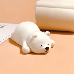 May include: A white ceramic polar bear figurine lying on its belly with its head turned to the side. The bear has black eyes and a black nose.