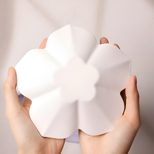 以下が含まれることがあります： 5つの花びらを持つ白いセラミックの花の形をした皿。皿は両手で持たれています