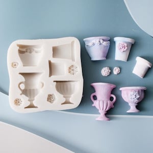 May include: A white silicone mold with various vessel and floral shapes alongside several finished light purple and pink miniature urns, pots, and flower embellishments cast from the mold, displaying detailed relief textures and classic forms.