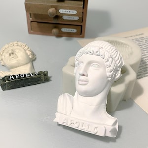 May include: Two white plaster busts of the Greek god Apollo. The busts are on a white surface. The bust on the left has a black base with the word "APOLLO" written on it. The bust on the right has a white base with the word "APOLLO" written on it.