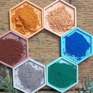 May include: Six hexagonal containers filled with different colored powders. The powders are shades of orange, brown, blue, gray, and green. The containers are arranged in a circle on a wooden surface.