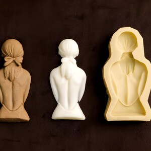 Peut inclure: Trois éléments sont présentés sur un fond marron foncé. À gauche, une sculpture de dos de personne couleur beige avec une queue de cheval. Au centre, une sculpture blanche d'un dos. À droite, un moule crème d'un dos.