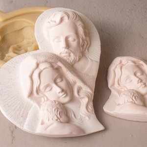 May include: A beige silicone heart-shaped mold featuring a relief of the Holy Family, alongside two white plaster castings. One casting depicts the Holy Family together, while the smaller casting shows the Virgin Mary holding the infant Jesus.