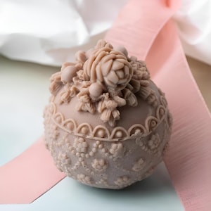 May include: A round, decorative soap with a floral design. The soap is a light brown colour and has a raised design of a rose and leaves. The soap is sitting on a white surface.