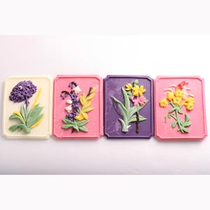 May include: Four square chocolate tiles featuring different floral designs. Each tile is a different colour: white, pink, purple, and pink. The flowers are made of chocolate and are in various shades of purple, yellow, and green.