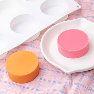 May include: A white silicone mould with four round cavities, a pink and an orange round soap on a pink and white checked surface.