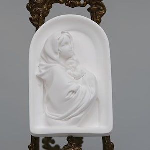 May include: A white relief sculpture of a woman holding a child, set within an arched frame. The sculpture is mounted on a decorative, ornate brass stand. The figures are draped in flowing garments.
