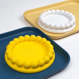 May include: Two decorative trays. One is a deep blue square tray with a bright yellow circular insert, edged with yellow spheres. The other is a yellow square tray with a white circular insert, edged with white spheres.