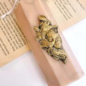 May include: A decorative bookmark with a gold-colored, ornate mask design featuring feathers and a face. The bookmark is set against a pale pink and beige background, with a book visible in the background.