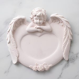 May include: A pale pink decorative tray shaped like a heart, featuring a sculpted cherub figure resting its arms at the top center. The tray is framed by two large, detailed feathered wings on either side and three small roses at the bottom base.