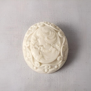 May include: Ivory-colored oval cameo with a detailed relief of a woman in profile, surrounded by floral and scrollwork designs. The cameo is a classic piece of jewelry or decorative art.