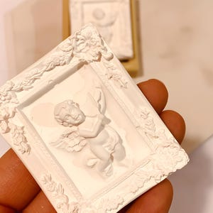 May include: White plaster cherub sculpture in a white ornate frame. The cherub is holding a scroll and has wings.