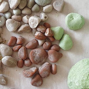 May include: A collection of smooth, rounded stones in various shades of earthy brown, muted gray, and pale green. The stones vary in size and texture, with some displaying natural pitting and color variations across their surfaces, scattered together.