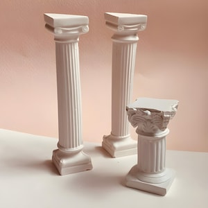 May include: Three white decorative columns of varying heights. Two tall, slender columns feature fluted shafts and simple capitals, while the third, shorter column has a wider, ornate capital with detailed carvings and a square base.