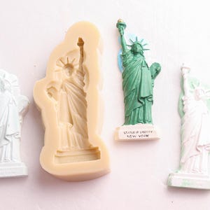 May include: A collection of Statue of Liberty figurines and a mold. The figurines are in white, green, and a light pink color. The mold is beige. The green statue has the text "STATUE OF LIBERTY NEW YORK" on the base.