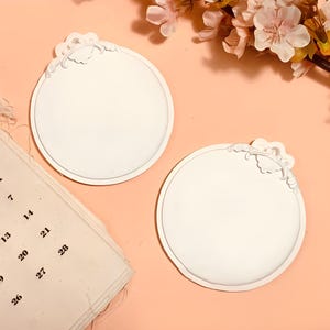 May include: Two white, round decorative plaques with a floral design on a peach background. A calendar is in the lower left corner, and pink flowers are in the upper right corner. The calendar shows the days of the week and the numbers 5, 6, 7, 12, 13, 14, 19, 20, 21, 26, 27, and 28.