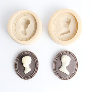 May include: Two silicone moulds for making chocolate or fondant cameo shapes. The moulds are beige and feature a raised design of a woman's profile in an oval frame.