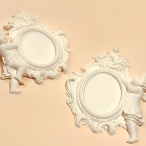 May include: Two white oval picture frames featuring sculpted cherub figures on the sides. Each frame has an ornate, raised border with floral and scroll details. The cherubs are depicted with wings and draped fabric, integrated into the frame design.