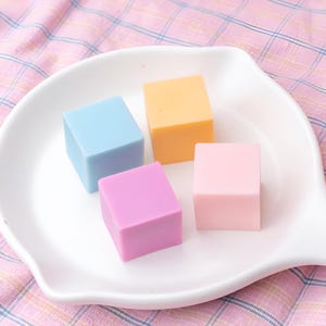 May include: Four small square blocks of soap in different colours: light blue, light purple, light pink, and light yellow. The blocks are arranged on a white plate.