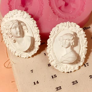 May include: Two white ceramic molds of ornate frames with a bust of a woman in each. The frames are detailed with intricate scrollwork and the busts are of a woman with long hair and a simple dress.