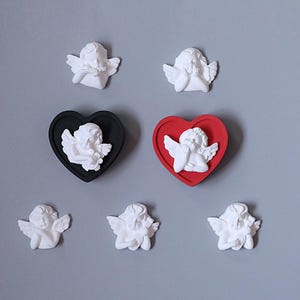 May include: A collection of seven white cherub figurines, with two of the cherubs mounted inside black and red heart-shaped frames. The cherubs are depicted in various poses, including resting their heads on their hands or holding small objects.