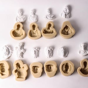 May include: A set of 14 silicone molds for making cake decorations. The molds are in the shape of various busts and figures, including a cherub, a lion, and a woman's head. The molds are beige in color and are arranged in two rows on a white surface.