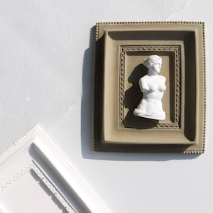 May include: A white plaster statue of a woman's torso in a brown frame with a decorative border.
