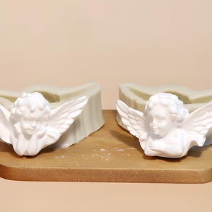 May include: Two white silicone moulds featuring a detailed cherub angel with outstretched wings. The moulds are designed for casting, showing the negative space of the angel figure in a soft, flexible material with a smooth finish.